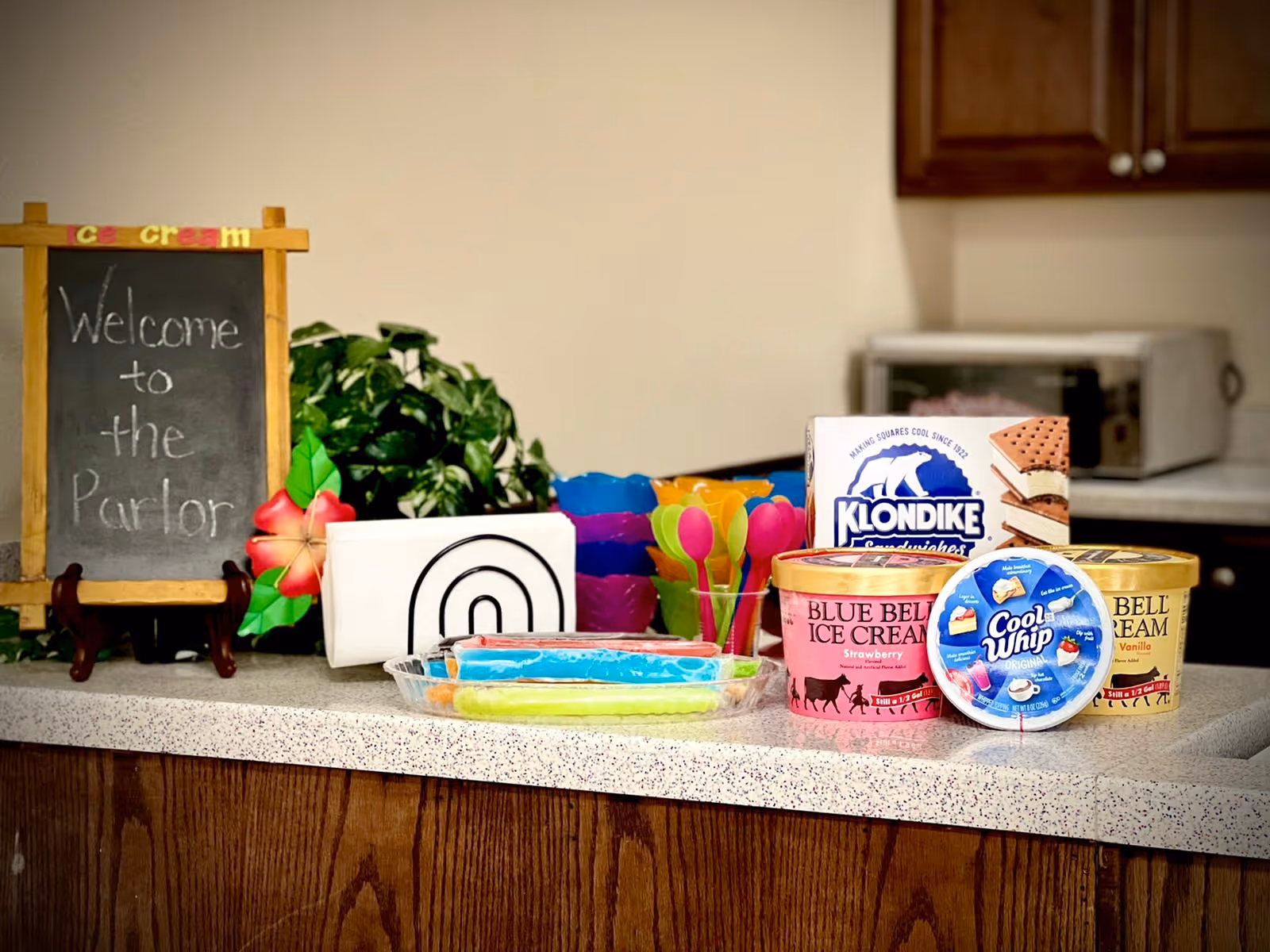Countertop with ice cream treats including Blue Bell ice cream, Klondike sandwiches, Cool Whip, colorful plastic spoons, and popsicles. A small chalkboard sign reads 'Welcome to the Parlor' with a decorative flower and a green plant in the background.