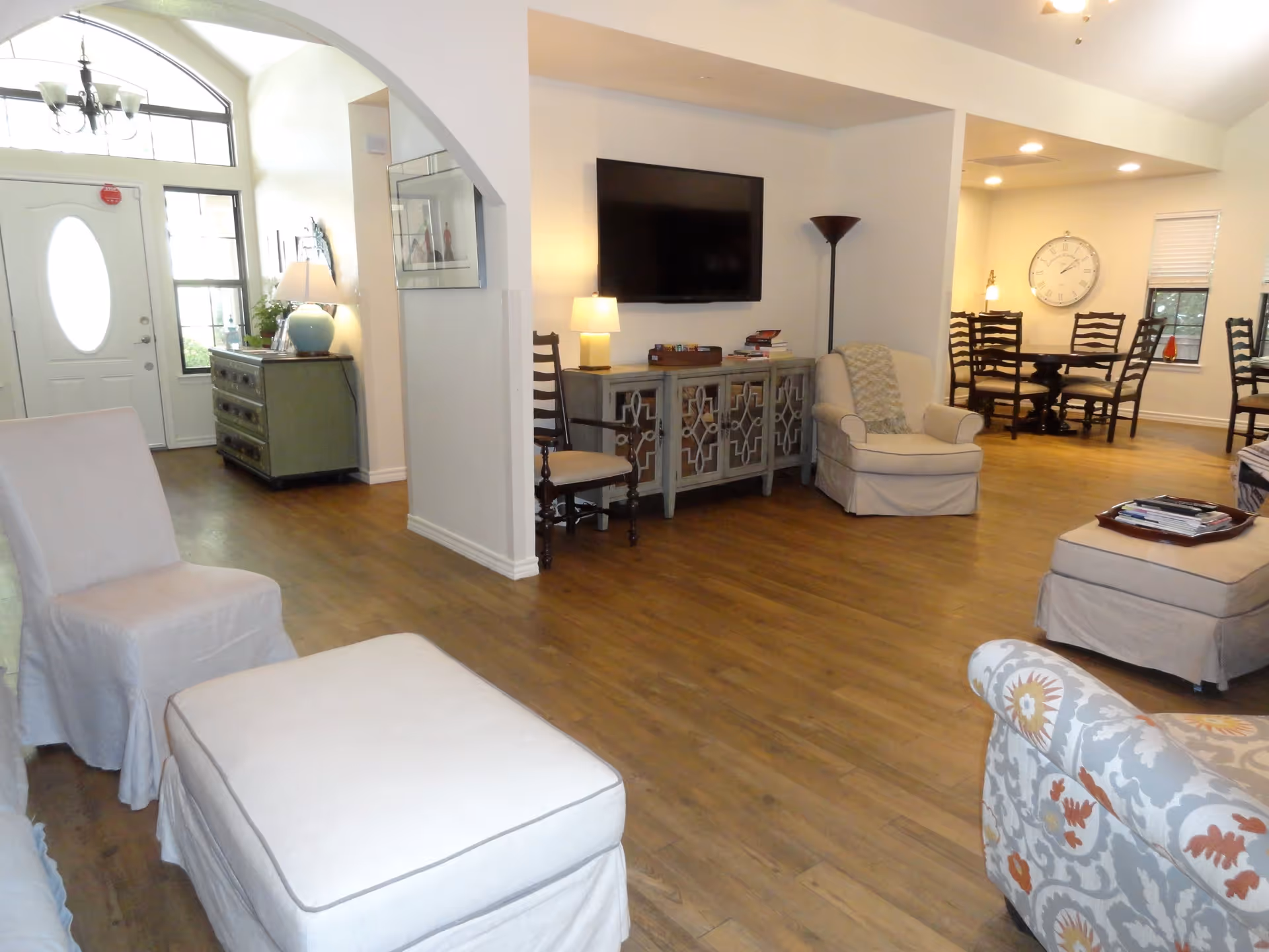 A spacious living room area with wooden flooring, featuring a white armchair with a matching ottoman, a patterned armchair, a TV mounted on the wall above a decorative cabinet, and a dining area with a round table and chairs in the background. The room is well-lit with lamps and natural light coming through the front door with glass panels.