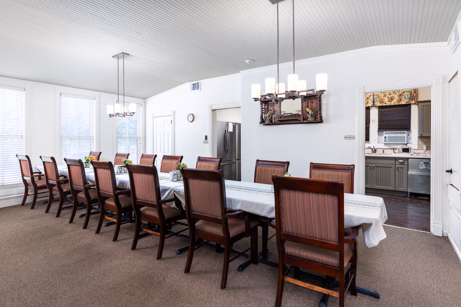 A long dining table covered with a white tablecloth and surrounded by wooden chairs with upholstered seats and backs in a bright dining room. The room has large windows with white blinds, two modern chandeliers hanging from a white paneled ceiling, and a doorway leading to a kitchen area with visible appliances and cabinetry.
