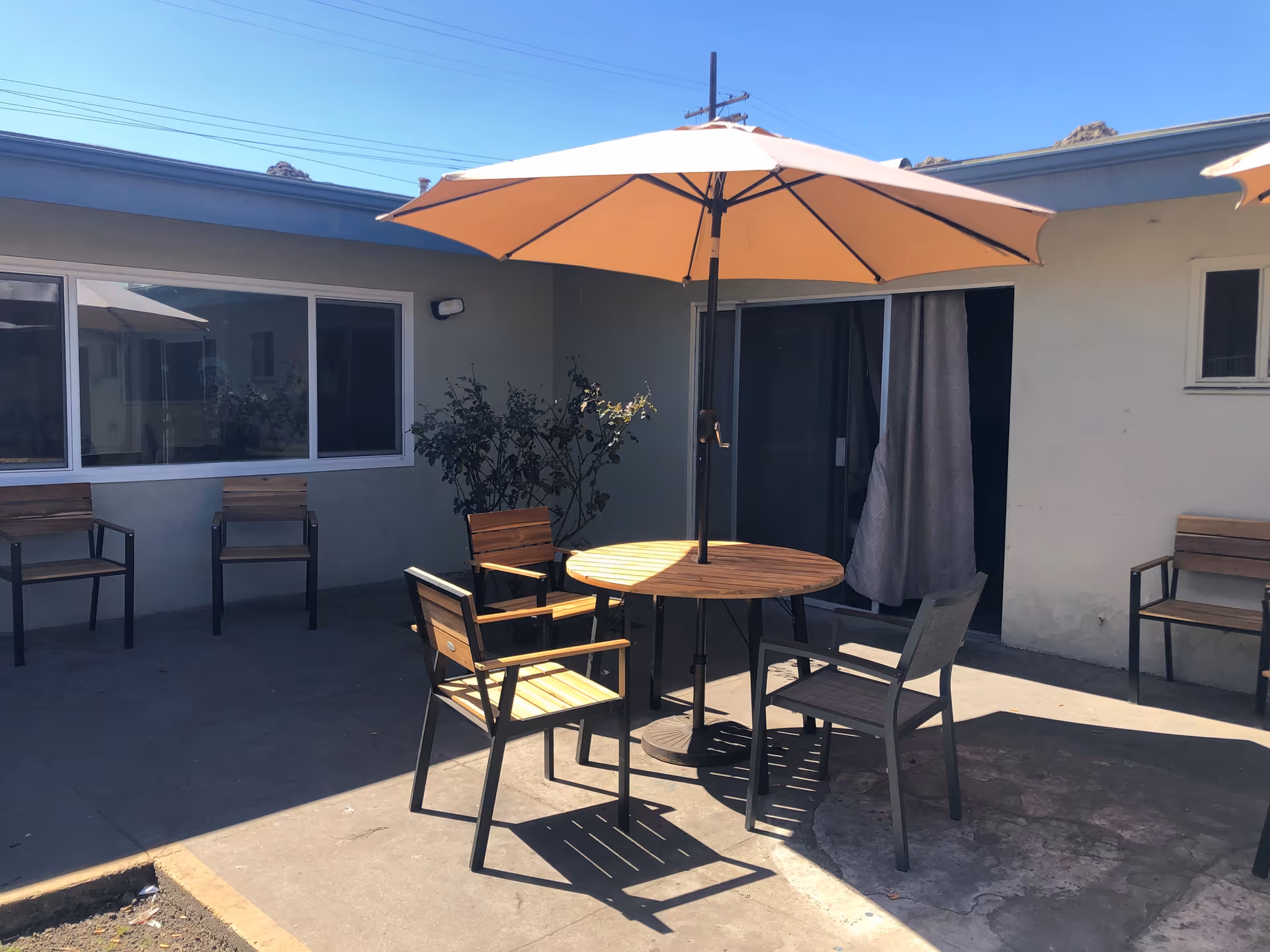 Outdoor patio area with a round wooden table and four chairs under a large beige umbrella. Additional wooden chairs are placed against the building wall. The building has sliding glass doors and windows, and there are some plants near the wall.
