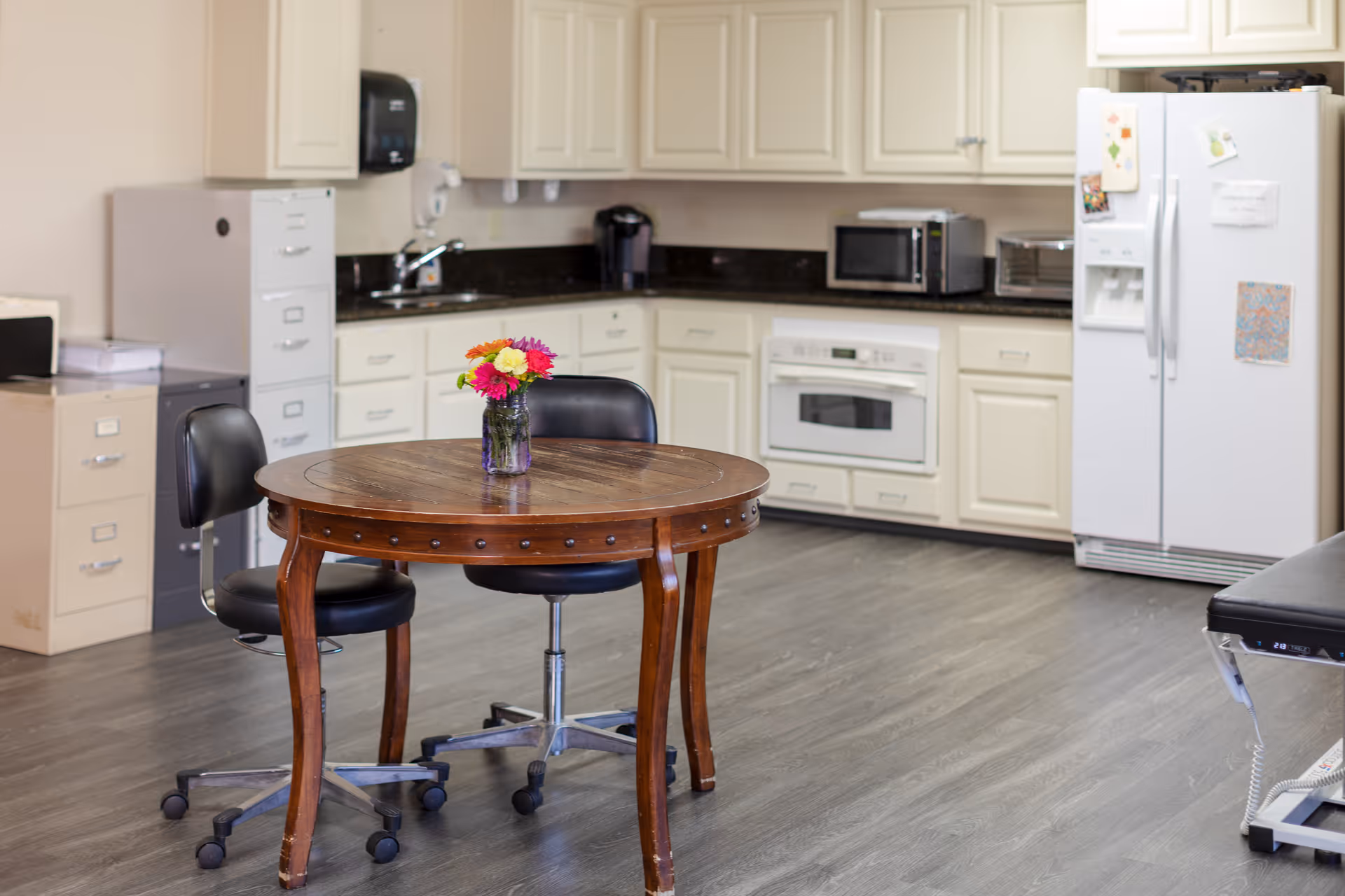 Communal kitchen area with a round wooden table and vase of flowers, two rolling chairs, white cabinets, microwave, and refrigerator.