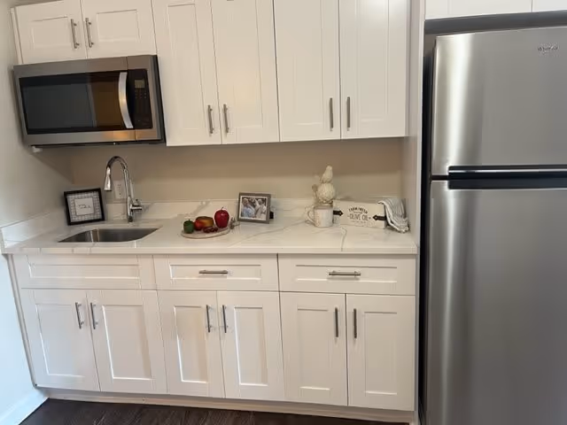 A modern kitchen area with white cabinets, a stainless steel microwave mounted above a sink, a stainless steel refrigerator, and a white countertop decorated with a small framed picture, a plate with fruits, and a decorative sign.