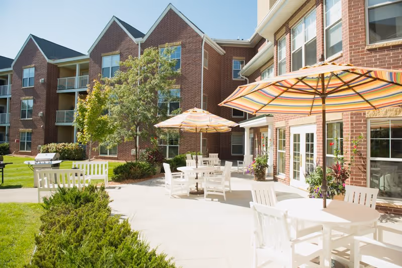 Outdoor patio area at The Waters of Eden Prairie featuring white tables and chairs with colorful striped umbrellas, surrounded by greenery and a brick building with multiple windows and balconies in the background under a clear blue sky.