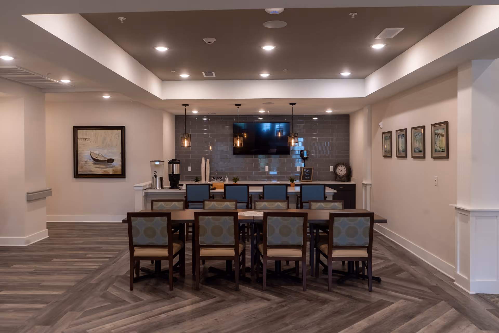 A modern dining area with a long wooden table surrounded by eight chairs with patterned upholstery. Behind the table is a counter with four high chairs, pendant lights hanging above, a large flat-screen TV mounted on a gray tiled wall, and various decorative items including a clock and framed pictures on the walls.