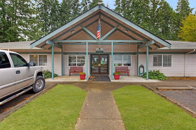 Front entrance of single-story memory care facility with a gabled porch, benches, and a parked pickup.