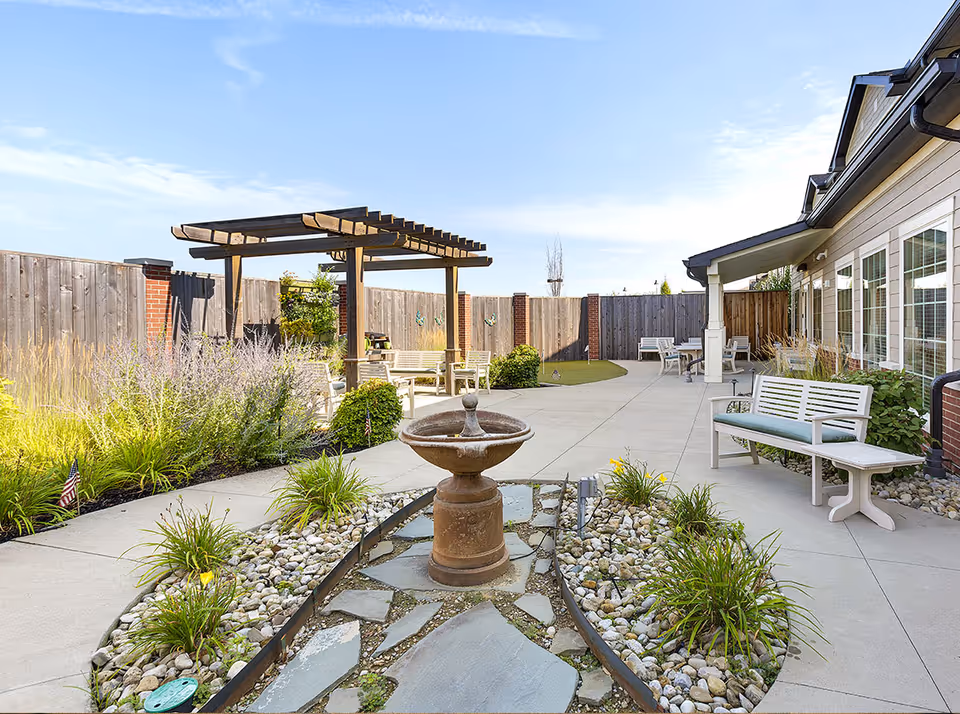 Outdoor courtyard area with a central stone water fountain surrounded by plants and rocks. There are benches with cushions, a wooden pergola with seating underneath, and a wooden fence enclosing the space. The sky is clear and blue.
