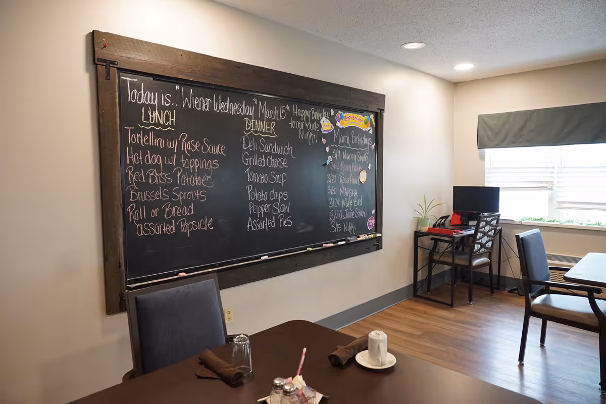 Interior view of a dining area in a senior living facility with a large chalkboard on the wall displaying the day's lunch and dinner menu, birthdays, and a birthday greeting. The room has tables and chairs, a small desk with a computer, a window with blinds, and wooden flooring.