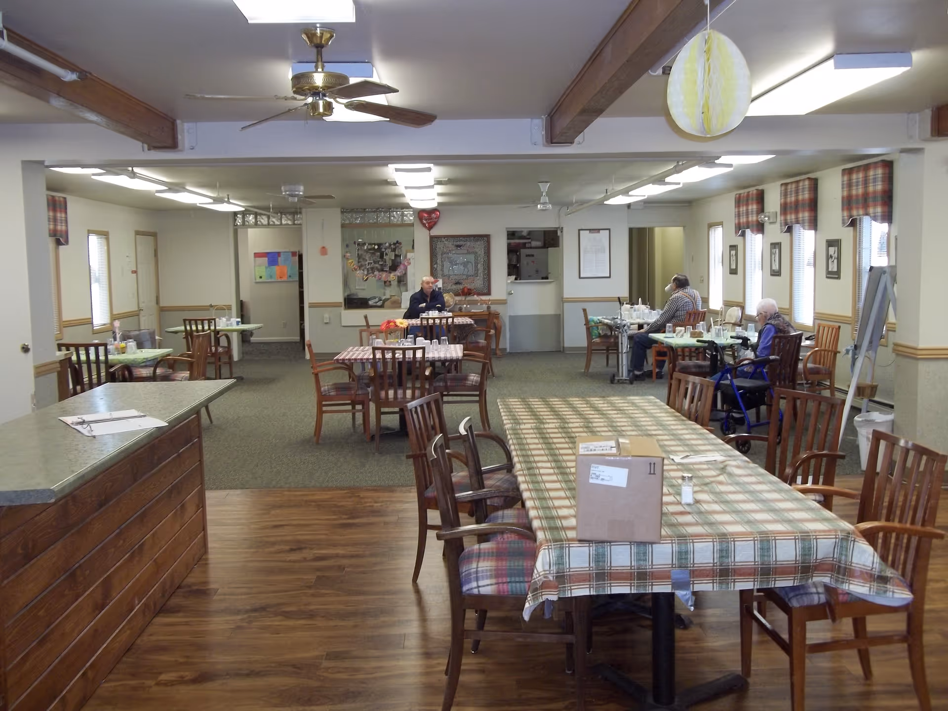 Open dining room in an assisted living community with long plaid-covered tables, wooden chairs, a serving counter, and a few residents seated.