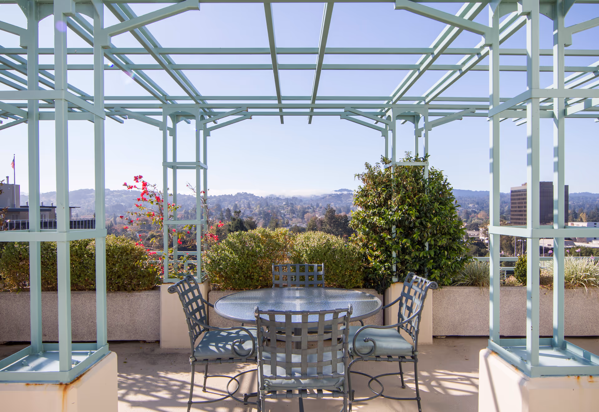 Rooftop terrace with a pale-green pergola, round glass table and four metal chairs overlooking planters and a distant hilly cityscape.