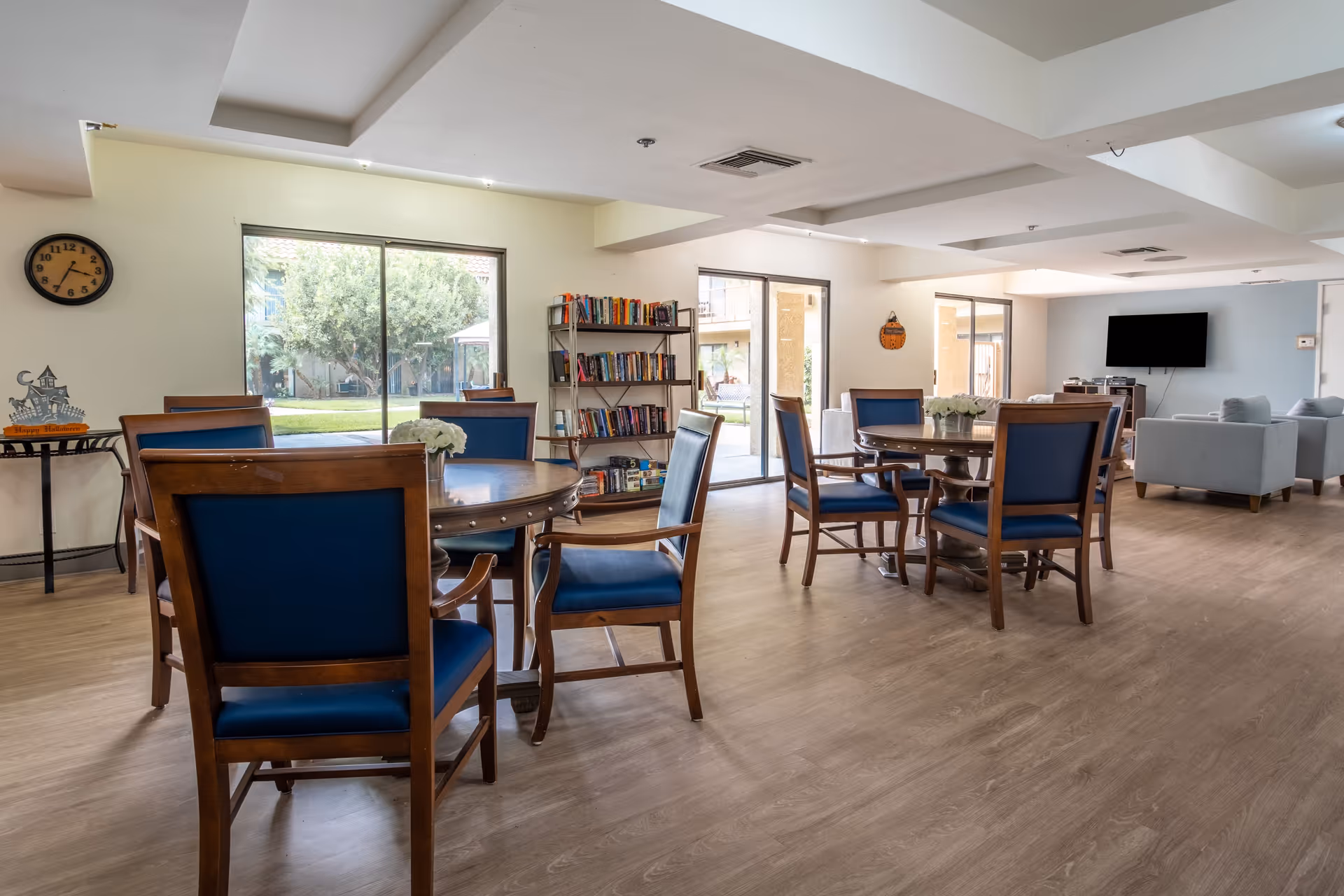 A spacious and well-lit common area in an assisted living facility featuring round wooden tables with blue cushioned chairs, a bookshelf filled with books, large windows and sliding glass doors showing a green outdoor area, a wall clock, and a seating area with gray sofas and a mounted flat-screen TV.