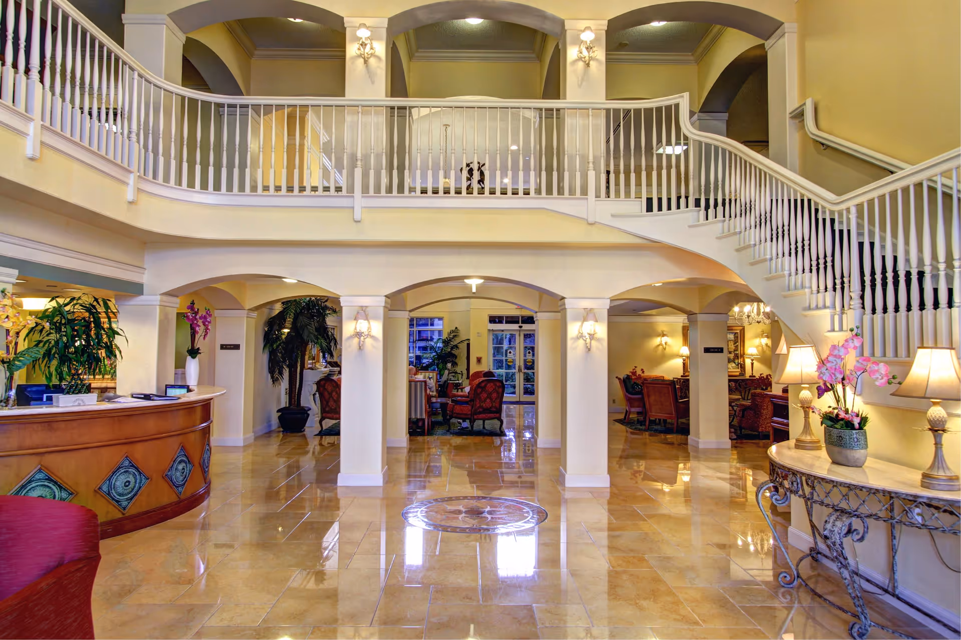 Spacious and elegant lobby area of a senior living facility featuring a polished tile floor, a curved staircase with white railings, decorative columns, and comfortable seating areas with armchairs. The space is warmly lit with wall sconces and table lamps, and decorated with plants and flowers.
