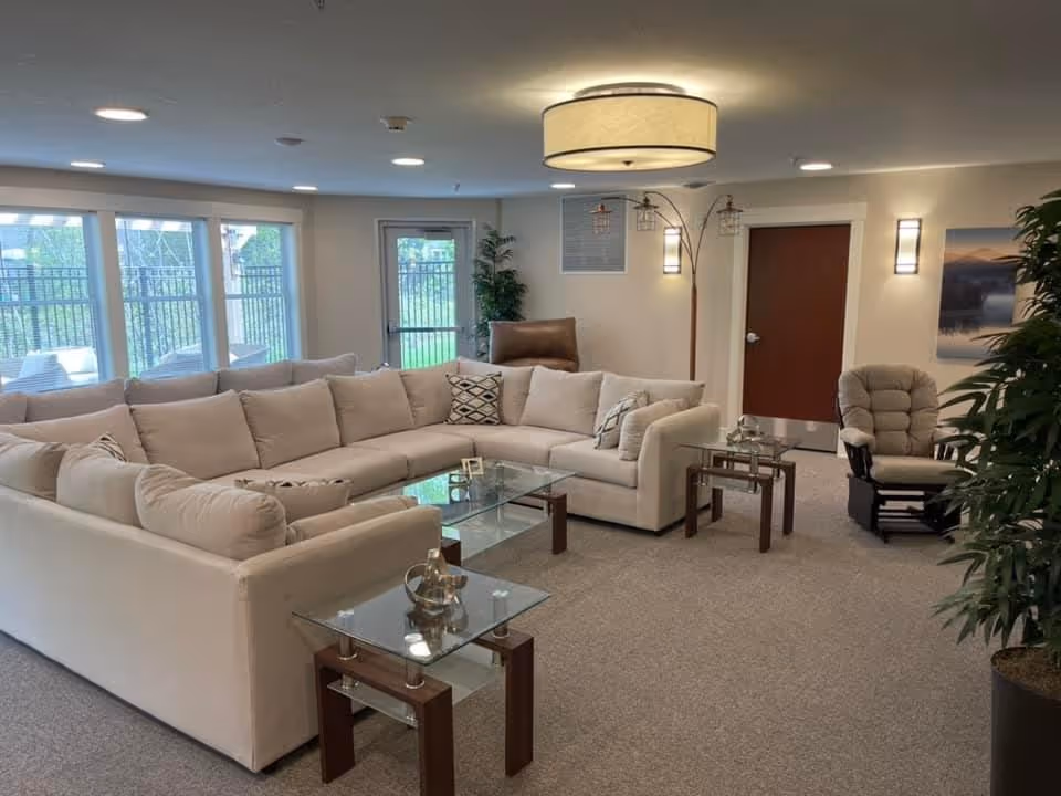 A spacious living room with a large beige sectional sofa arranged around a glass coffee table. There are two glass side tables with wooden legs, a cushioned rocking chair, and a tall floor lamp with three lights. The room has beige walls, carpeted floor, and several windows letting in natural light. A door and some green plants are also visible.