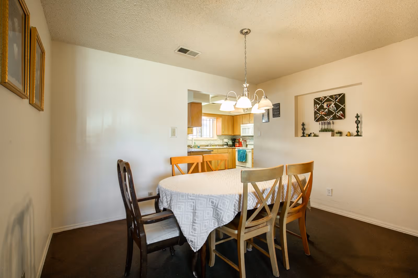A dining room with a round table covered by a white patterned tablecloth and surrounded by five wooden chairs. A chandelier with five lights hangs above the table. The room has beige walls and carpeted floor. There is a small open wall niche with decorative items and a view into a kitchen with wooden cabinets and appliances.