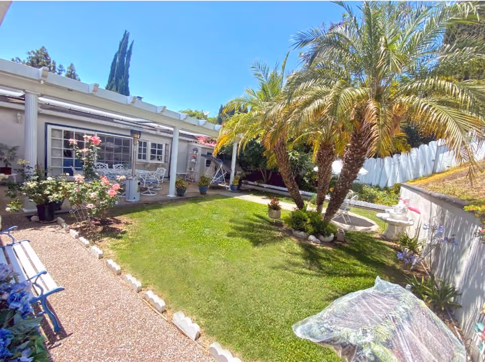 A sunny outdoor garden area with a well-maintained green lawn, palm trees, flowering plants, and a white fence. There is a covered patio with white chairs and a table, and a pathway lined with flowers and a bench on the left side.