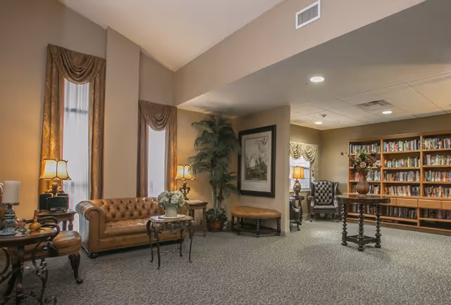 A cozy living room area with a brown leather tufted sofa, two tall windows with gold curtains, side tables with lamps, a round table with a flower vase, a large potted plant, framed artwork on the wall, and a bookshelf filled with books in the background.