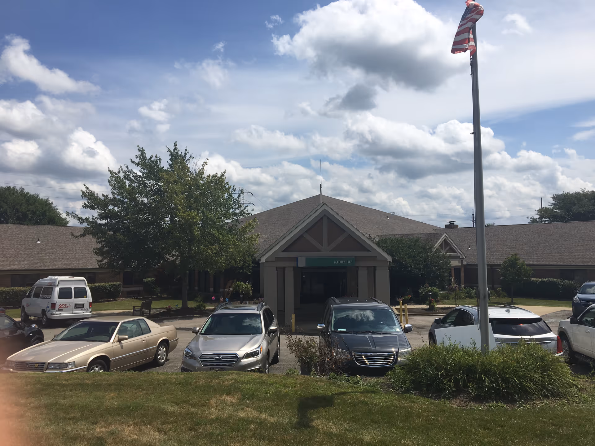 Front exterior view of Beeghly Oaks Center for Rehabilitation & Healing building with a parking lot in front, several parked cars, a flagpole with an American flag, and trees under a partly cloudy sky.