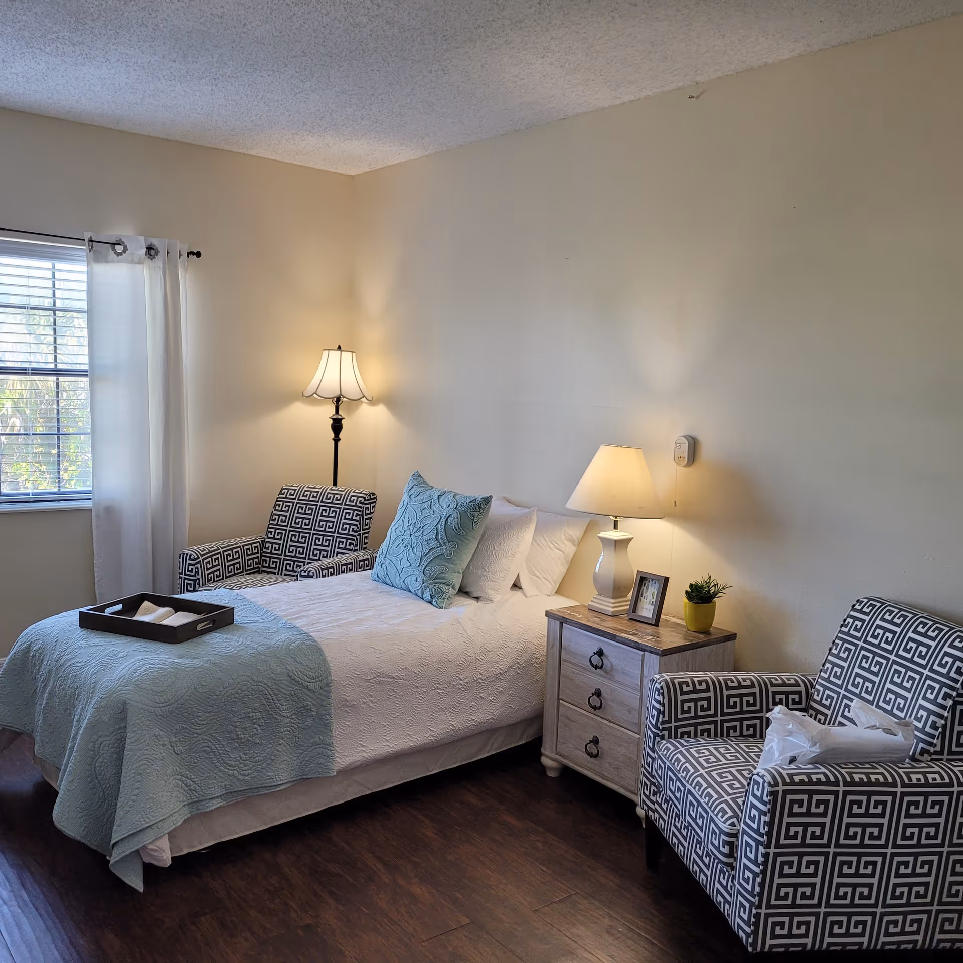 A cozy bedroom with a bed covered in white and light blue bedding, flanked by two patterned armchairs. A wooden nightstand with a lamp, small plant, and framed photo sits next to the bed. A floor lamp stands in the corner near a window with white curtains, and the room has wooden flooring and beige walls.