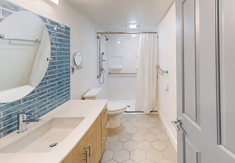 A modern bathroom featuring a white countertop with a built-in sink and wooden cabinets below. Above the sink is a round mirror mounted on a wall with blue subway tiles. The bathroom has a white toilet and a walk-in shower with a white curtain and grab bars. The floor is covered with hexagonal tiles, and the walls are painted white.