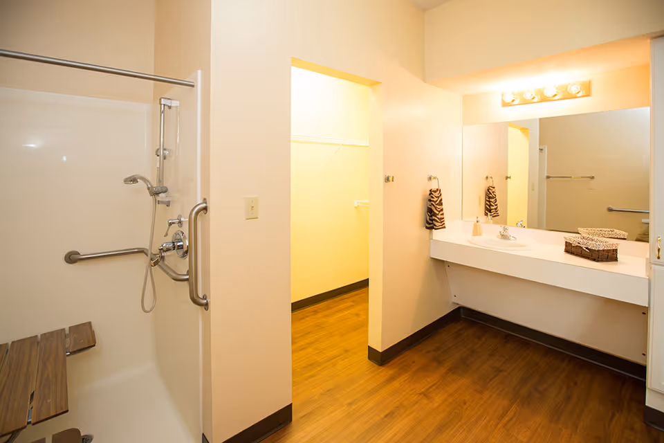 A bathroom with a walk-in shower featuring a handheld showerhead and safety grab bars. There is a wooden fold-down seat inside the shower. The room has a large mirror above a white sink countertop with a soap dispenser and a basket. The floor is wood-style, and there is an open doorway leading to a closet with shelving and hanging rods.