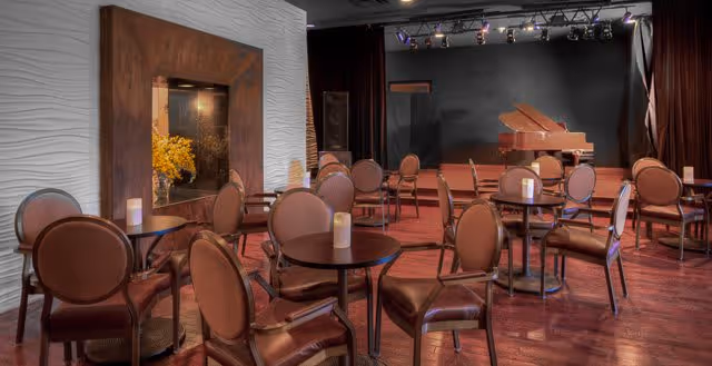 A cozy performance or event room with multiple round tables and brown cushioned chairs arranged on a wooden floor. There is a grand piano on a small stage with stage lighting above. A decorative fireplace with a wooden frame and yellow flowers inside is visible on the left side of the room.
