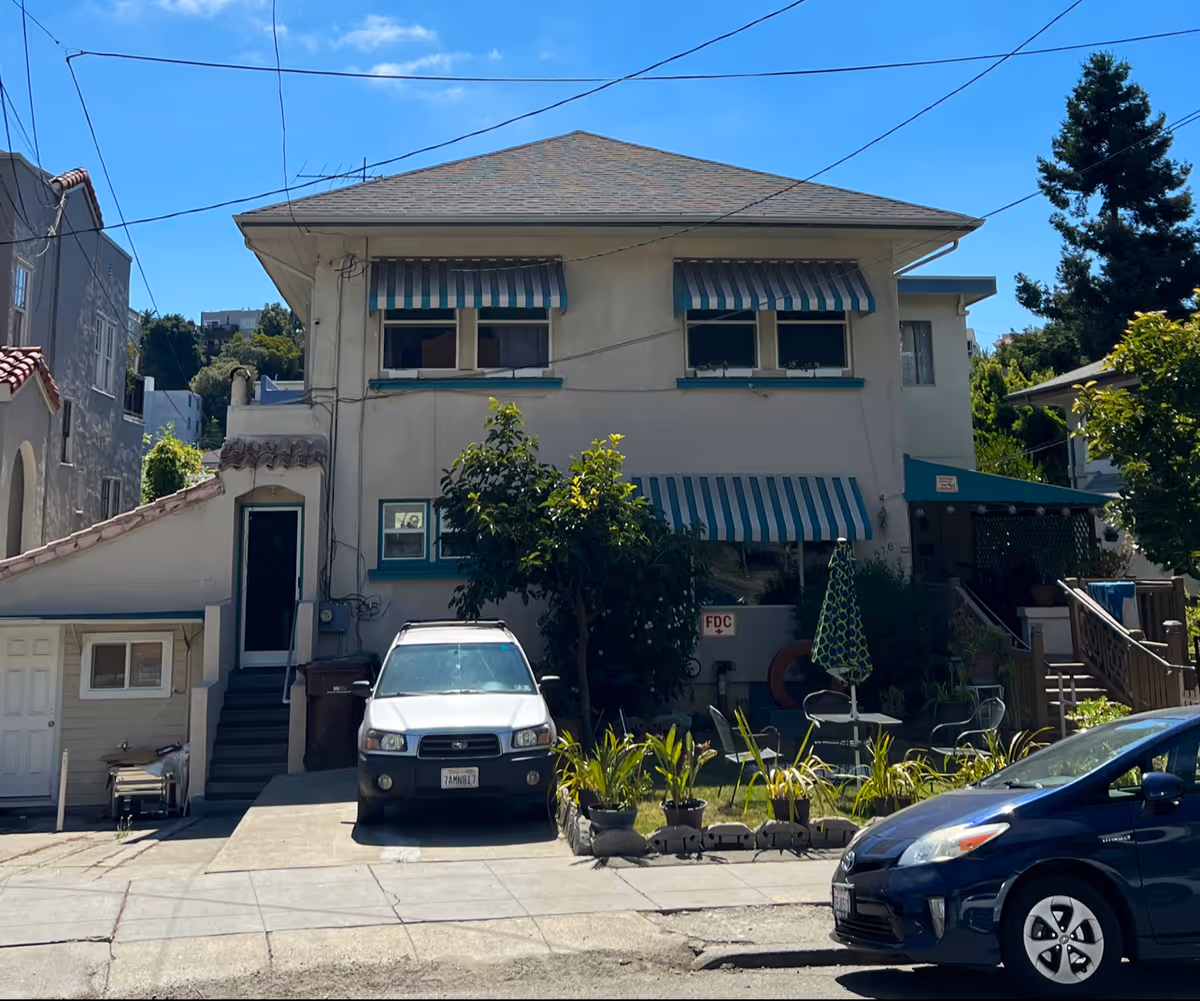 Exterior view of a two-story residential building with striped green and white awnings over the windows. There are two cars parked in front, a white SUV and a dark blue sedan. The building has a small garden area with plants and an umbrella-covered seating area. The sky is clear and blue.