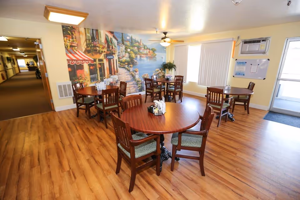 A dining area with several round wooden tables and chairs arranged on a wood floor. The walls are painted light yellow, and one wall features a large mural depicting a scenic outdoor cafe by the water with colorful buildings and flowers. There is a ceiling fan with a light fixture, a window with vertical blinds, an air conditioning unit, and a whiteboard on the wall. A hallway is visible to the left, and a glass door to the right leads outside.