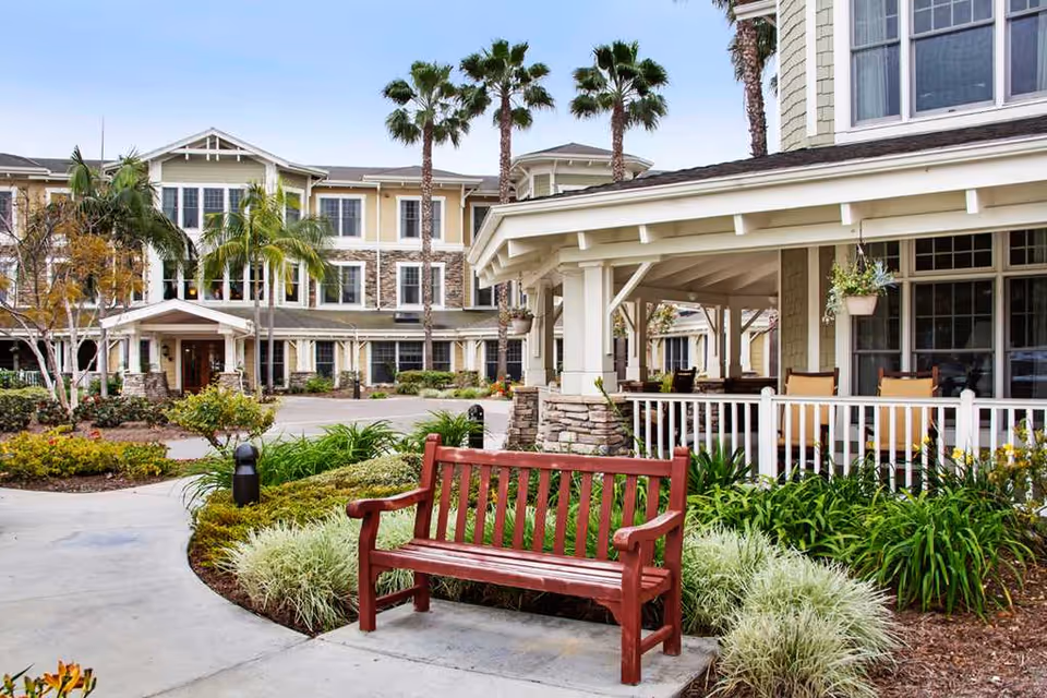 Outdoor area of a senior living facility featuring a red wooden bench on a concrete pathway surrounded by green plants and landscaping. In the background, there is a multi-story building with stone and beige siding, large windows, palm trees, and a covered porch with seating.