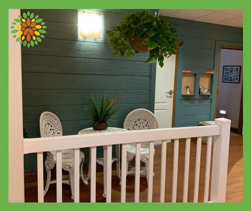 Indoor seating area with two white ornate metal chairs and a small round table with a plant on top. The background features a green paneled wall with a wall light and a hanging plant. There is a white railing in the foreground and a door and small shelves on the right side.