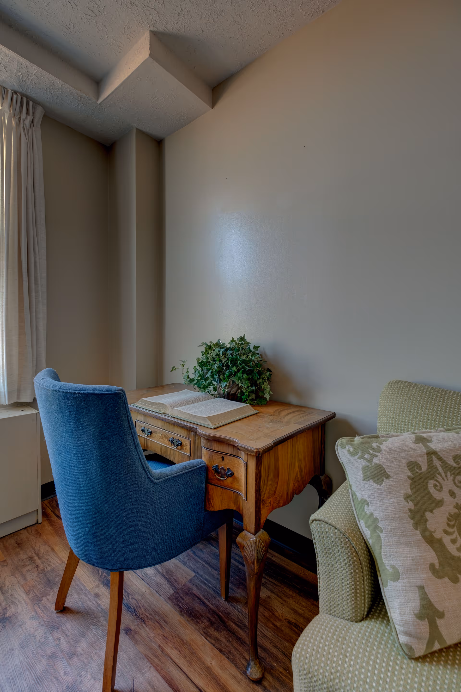 A cozy corner with a wooden desk holding an open book and a green potted plant. A blue upholstered chair is placed in front of the desk, and part of a green armchair with a patterned cushion is visible on the right. Light filters in through a window with cream-colored curtains on the left.