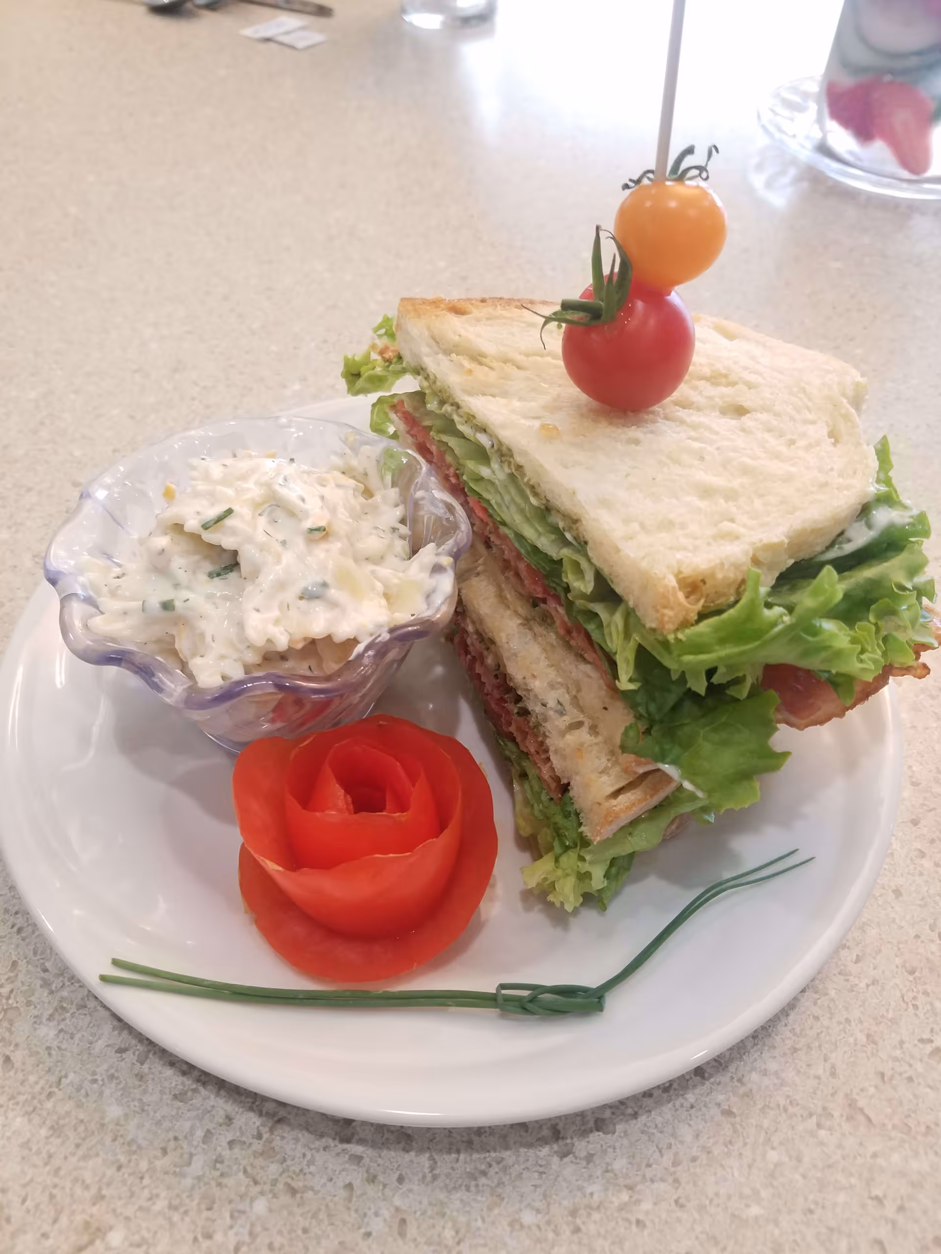 A plate with a club sandwich containing lettuce, tomato, and bacon, garnished with two cherry tomatoes on a toothpick. Next to the sandwich is a small glass bowl of creamy salad and a tomato carved into the shape of a rose, with a chive placed on the plate.