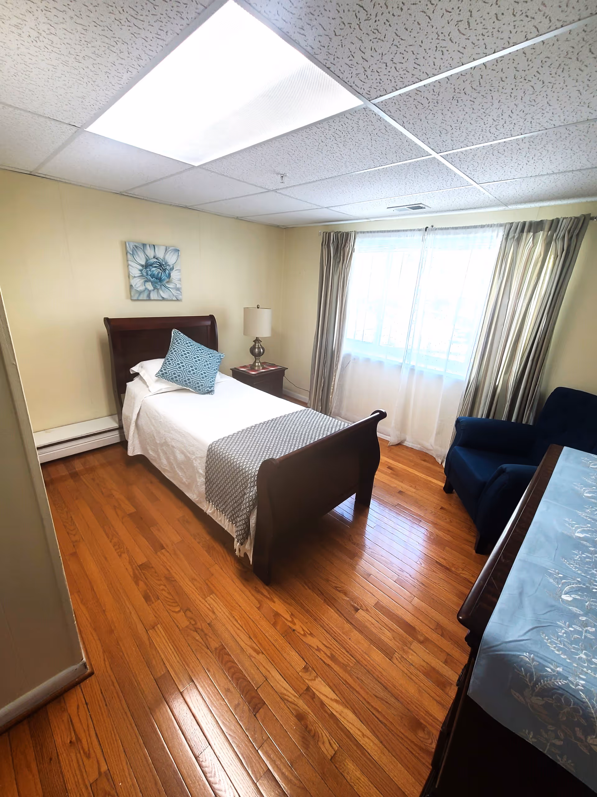 A small bedroom with a single wooden bed covered with white bedding and a blue patterned pillow. There is a wooden nightstand with a lamp next to the bed, a window with sheer and striped curtains, a blue armchair, and a wooden dresser with a blue decorative cloth on top. The floor is hardwood and the ceiling has white acoustic tiles with a fluorescent light panel.
