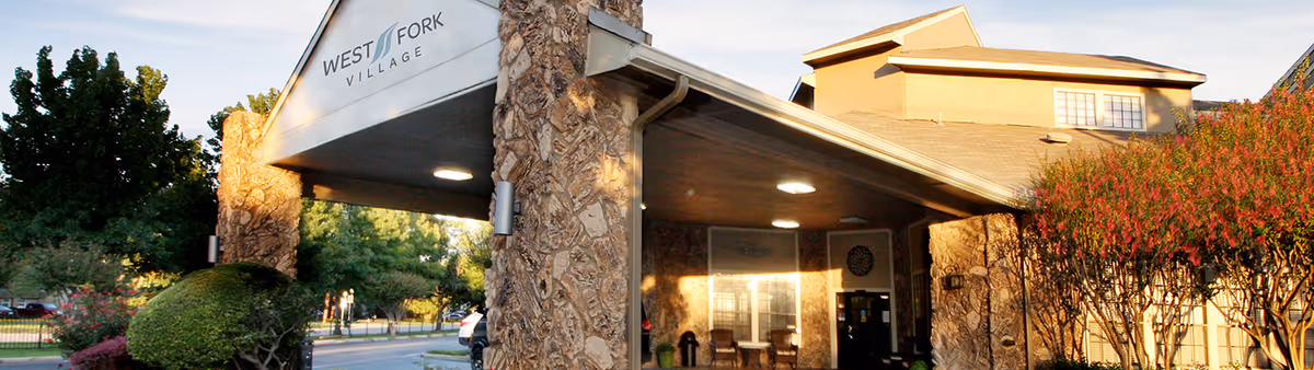 Entrance of West Fork Village facility with a covered driveway supported by stone pillars, surrounded by trees and bushes under a clear sky.