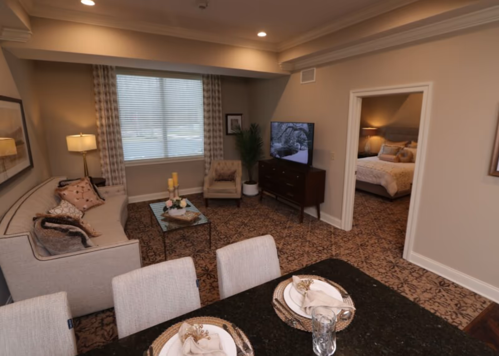 A cozy living room with a beige sofa adorned with decorative pillows, a glass coffee table with candles and flowers, an armchair, and a TV on a wooden stand. The room has patterned carpet and a large window with curtains. Adjacent to the living room is a bedroom visible through an open doorway, featuring a bed with pillows and a bedside lamp. In the foreground, there is a dining table set with plates, napkins, and glasses.
