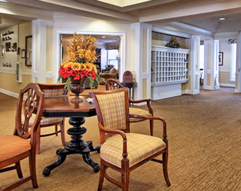 A spacious and well-lit interior common area with beige carpet and cream-colored walls. There is a round wooden table with a decorative vase of flowers in the center, surrounded by wooden chairs with plaid upholstery. The background shows a hallway with multiple doorways and a wall-mounted mail organizer.
