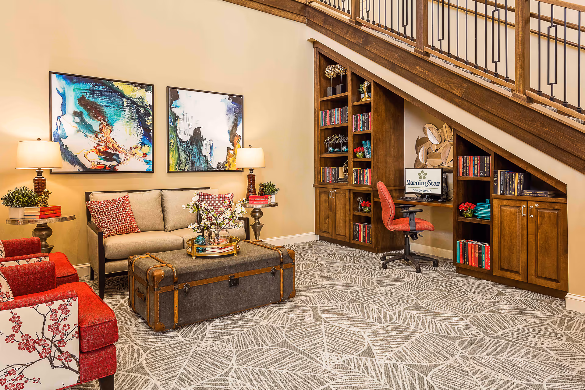 A cozy living room area with a beige sofa, two red armchairs, and a vintage trunk used as a coffee table. The walls are decorated with two abstract paintings. There are two side tables with lamps and plants. Under a wooden staircase, there is a built-in wooden bookshelf and a small desk with a red office chair and a computer monitor displaying the MorningStar logo.