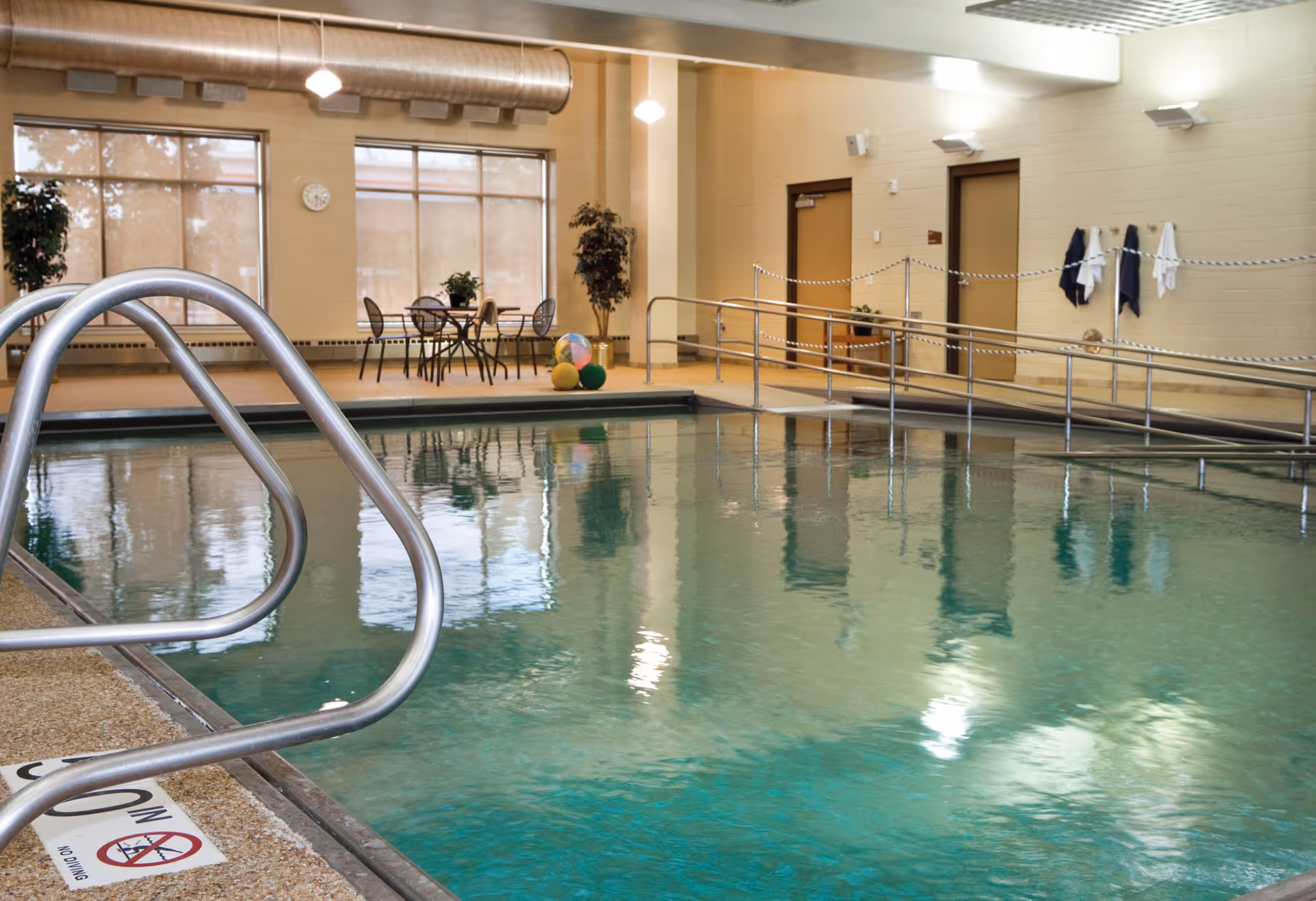 Indoor swimming pool with metal handrails, an accessibility ramp, and a small table-and-chairs seating area.