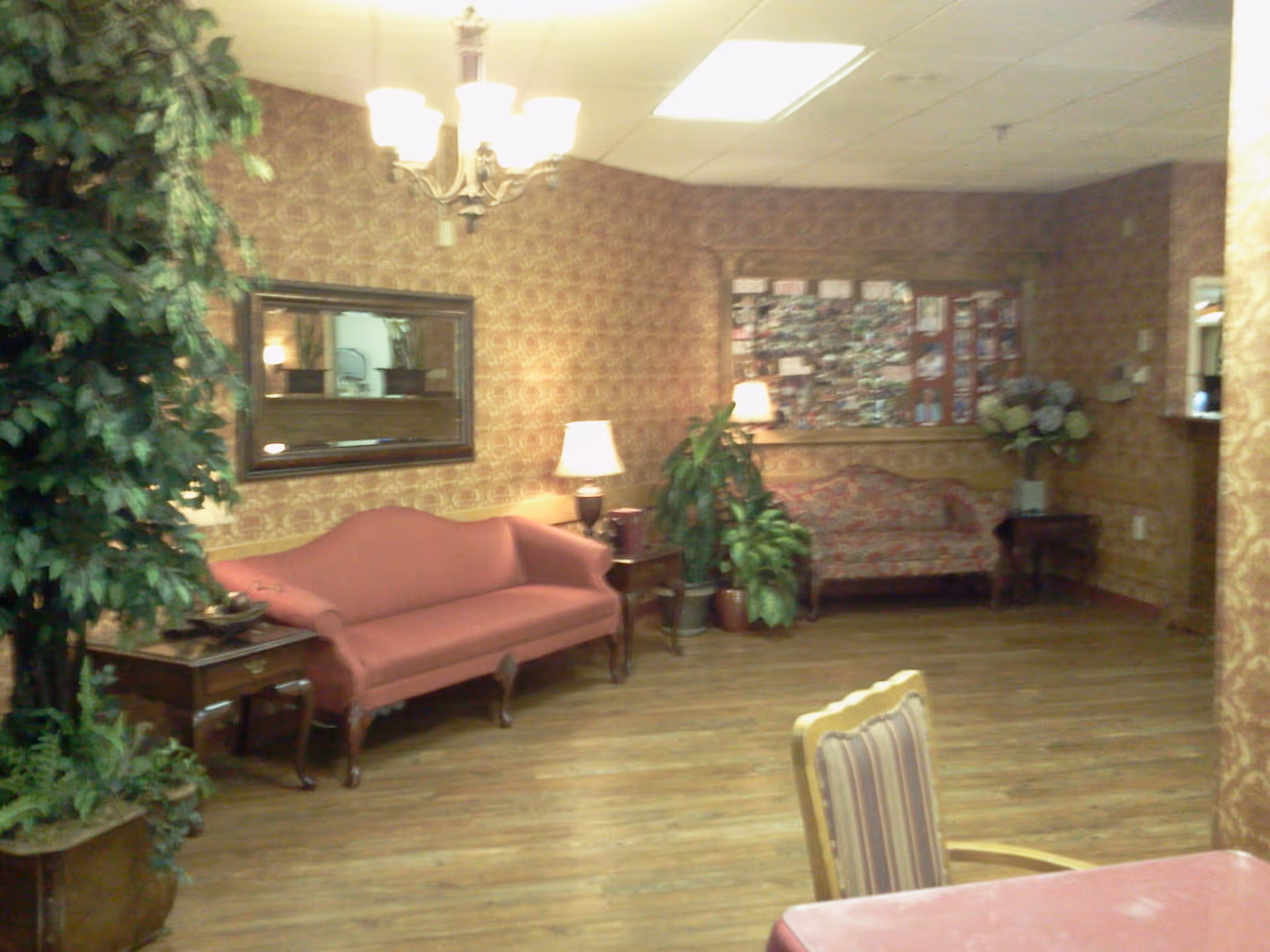 A cozy sitting area in a healthcare facility with two upholstered sofas, a wooden side table with a lamp, potted plants, a large mirror on the wall, and a bulletin board filled with photos and notes. The room has patterned wallpaper and wood flooring.
