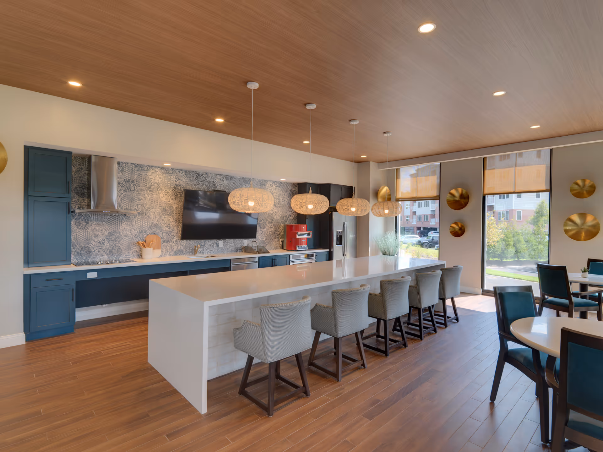 Modern communal kitchen and dining area with a long white island counter surrounded by six cushioned chairs. The kitchen features blue cabinets, a stainless steel refrigerator, a wall-mounted TV, and a decorative tiled backsplash. Pendant lights hang above the island, and large windows with shades allow natural light to fill the space. Additional dining tables and chairs are visible near the windows.