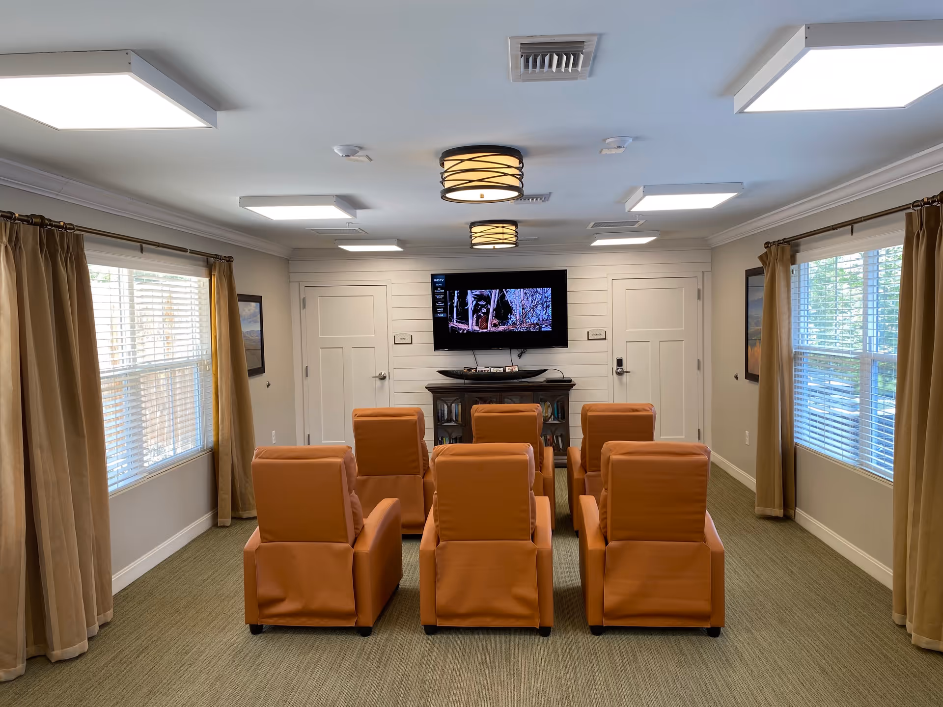 A small media room with six orange recliner chairs arranged in two rows facing a wall-mounted flat screen TV. The room has beige walls, two windows with beige curtains, and ceiling lights including two round fixtures and several rectangular panels. There are two closed white doors on either side of the TV.