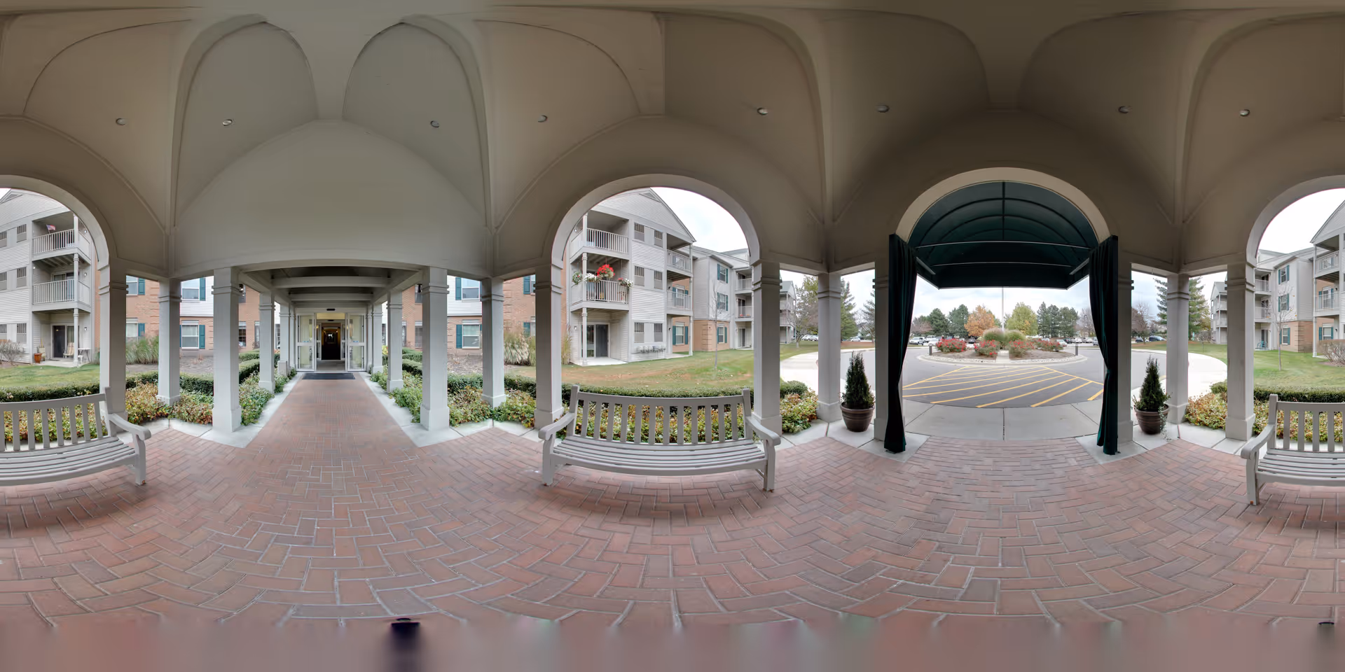 Covered entrance walkway of Oakmont Manor Senior Community with white benches on either side, brick flooring, white columns, and a green awning over the driveway. The building exterior is visible with balconies and windows, and there are landscaped areas with grass and shrubs.