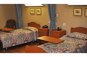 A healthcare facility bedroom with two single beds, each covered with patterned bedspreads. Between the beds is a wooden nightstand with a telephone and a lamp. There is a small table in front of the beds and two chairs near the window with blue curtains. The walls have framed artwork.