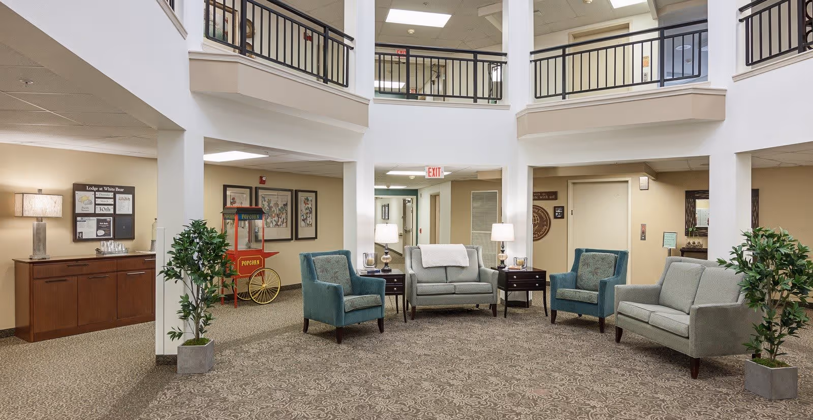 A spacious and well-lit common area in The Lodge at White Bear, featuring comfortable seating with two armchairs and two loveseats arranged around two side tables with lamps. The area has a patterned carpet, potted plants, and a popcorn machine in the background. The space is open to a second-floor balcony with black railings and has beige walls and ceiling lights.