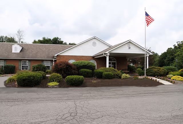 Front exterior view of a single-story brick building with a white gabled roof and a covered entrance. There are neatly trimmed bushes and landscaping in front of the building, and an American flag on a flagpole near the entrance. The sky is cloudy.