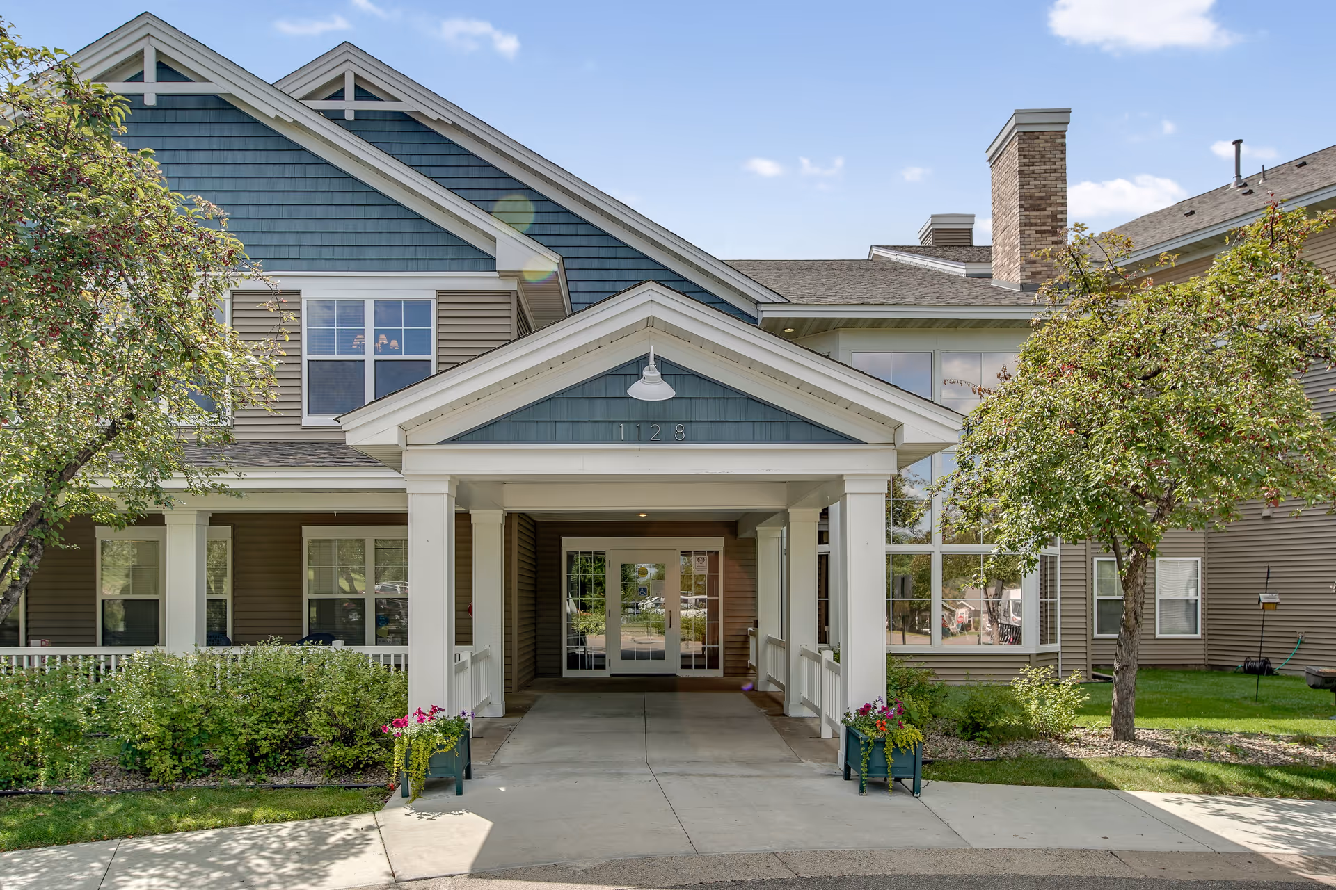 Front exterior view of Oak Ridge Assisted Living facility showing a covered entrance with white columns, a pathway leading to glass double doors, and surrounding greenery including trees and flower pots.