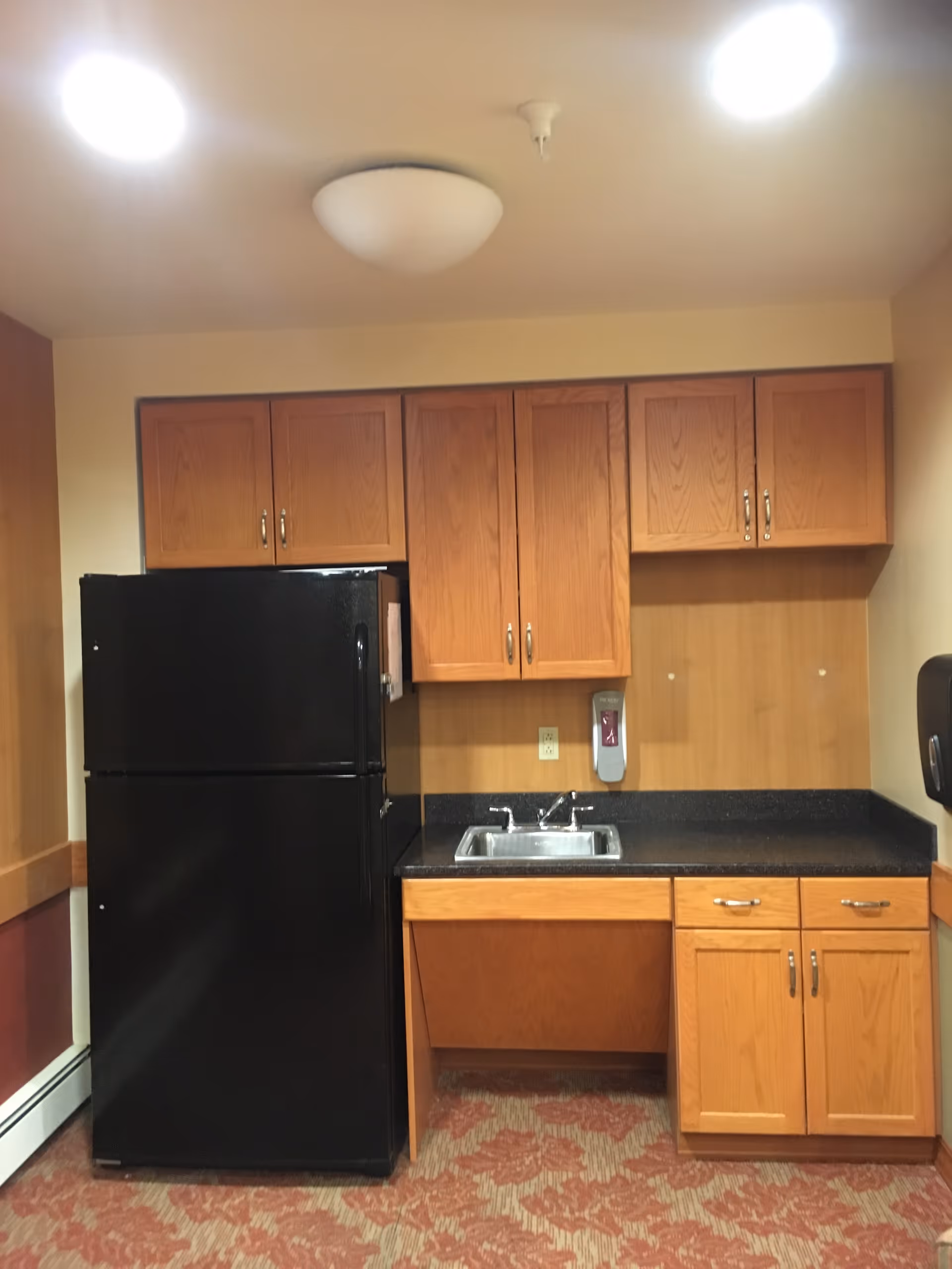 Kitchenette area with a black refrigerator, stainless steel sink, and oak cabinets over a dark countertop.