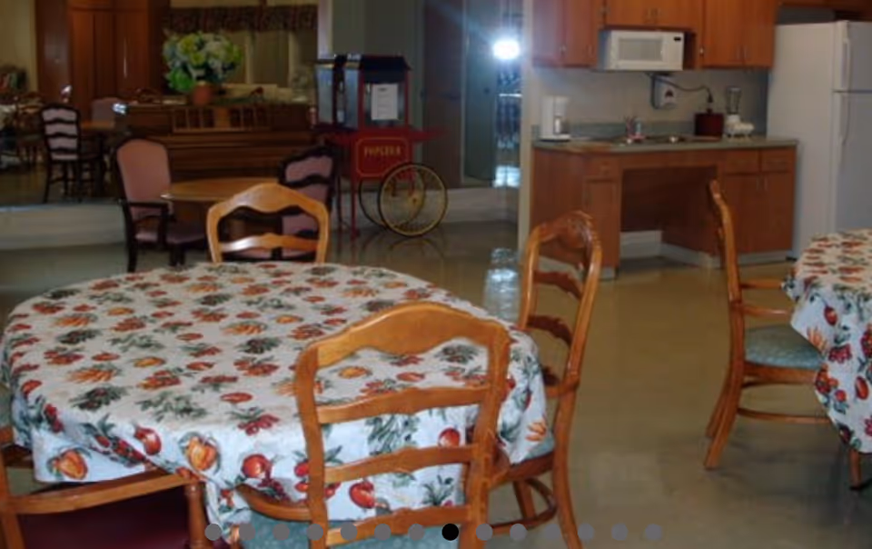 Interior view of a dining area with round tables covered in floral tablecloths and wooden chairs. In the background, there is a kitchenette with wooden cabinets, a microwave, a refrigerator, and a popcorn machine. The floor is tiled and the room has a cozy, home-like atmosphere.