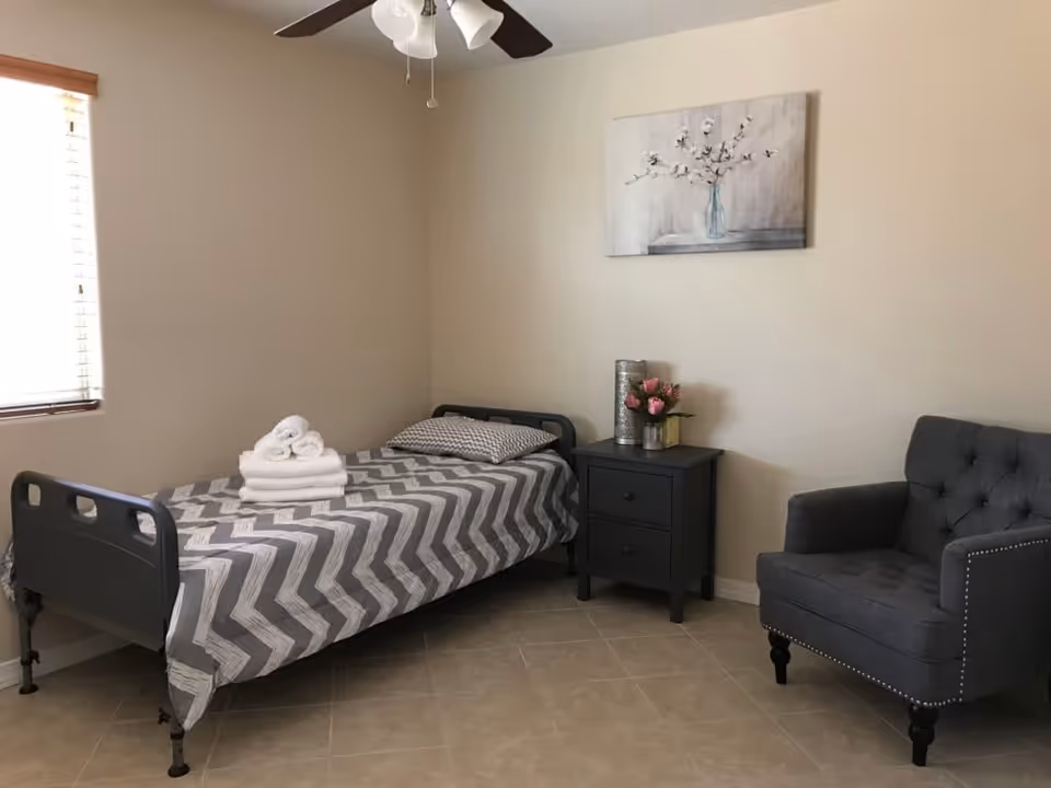 A simple bedroom with a single bed covered in a gray and white chevron patterned bedspread, a pillow, and a stack of folded white towels on top. Next to the bed is a black nightstand with two drawers, decorated with a vase of pink flowers and a cylindrical decorative item. A gray upholstered armchair with button tufting and nailhead trim is positioned to the right of the nightstand. The room has beige walls, a tiled floor, a window with blinds on the left, a ceiling fan with lights, and a painting of flowers in a vase hanging above the nightstand.