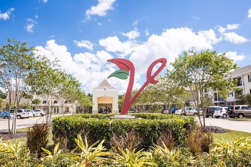 Front exterior of a senior living facility with a large red 'V' sculpture in a landscaped roundabout, surrounding buildings and parked cars under a blue sky.