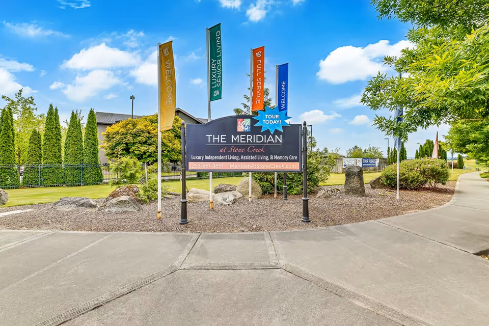 Outdoor view of the entrance area to The Meridian at Stone Creek senior living facility, featuring a large sign with the facility name and services offered, surrounded by colorful flags and landscaped greenery under a blue sky with clouds.