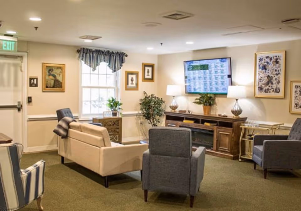 A cozy living room area in a senior living facility with a beige sofa, two gray armchairs, and a striped armchair arranged around a wooden entertainment console with a flat-screen TV mounted above it. The room has framed artwork on the walls, two table lamps on the console, a window with a valance, and some potted plants. There is an exit door on the left side of the room.
